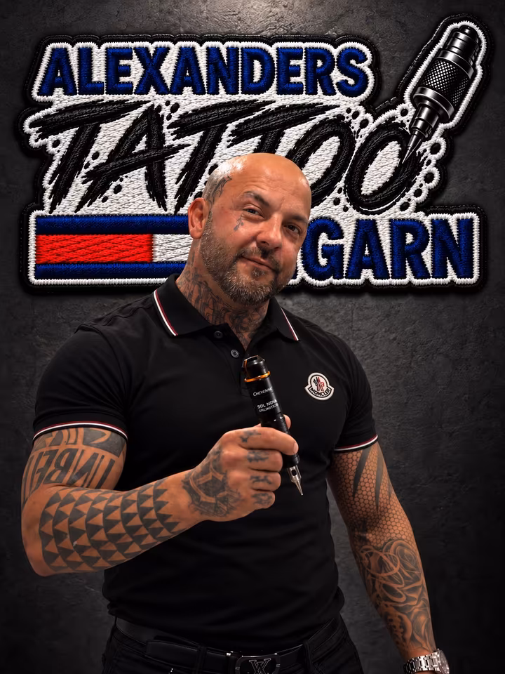 Alexander Garn mit Tattoo-Maschine in der Hand vor dem Logo seines Studios